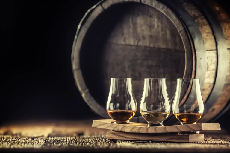 Glencairn whiskey tasting cups on a wooden serving, with a whisky barrel in the dark background